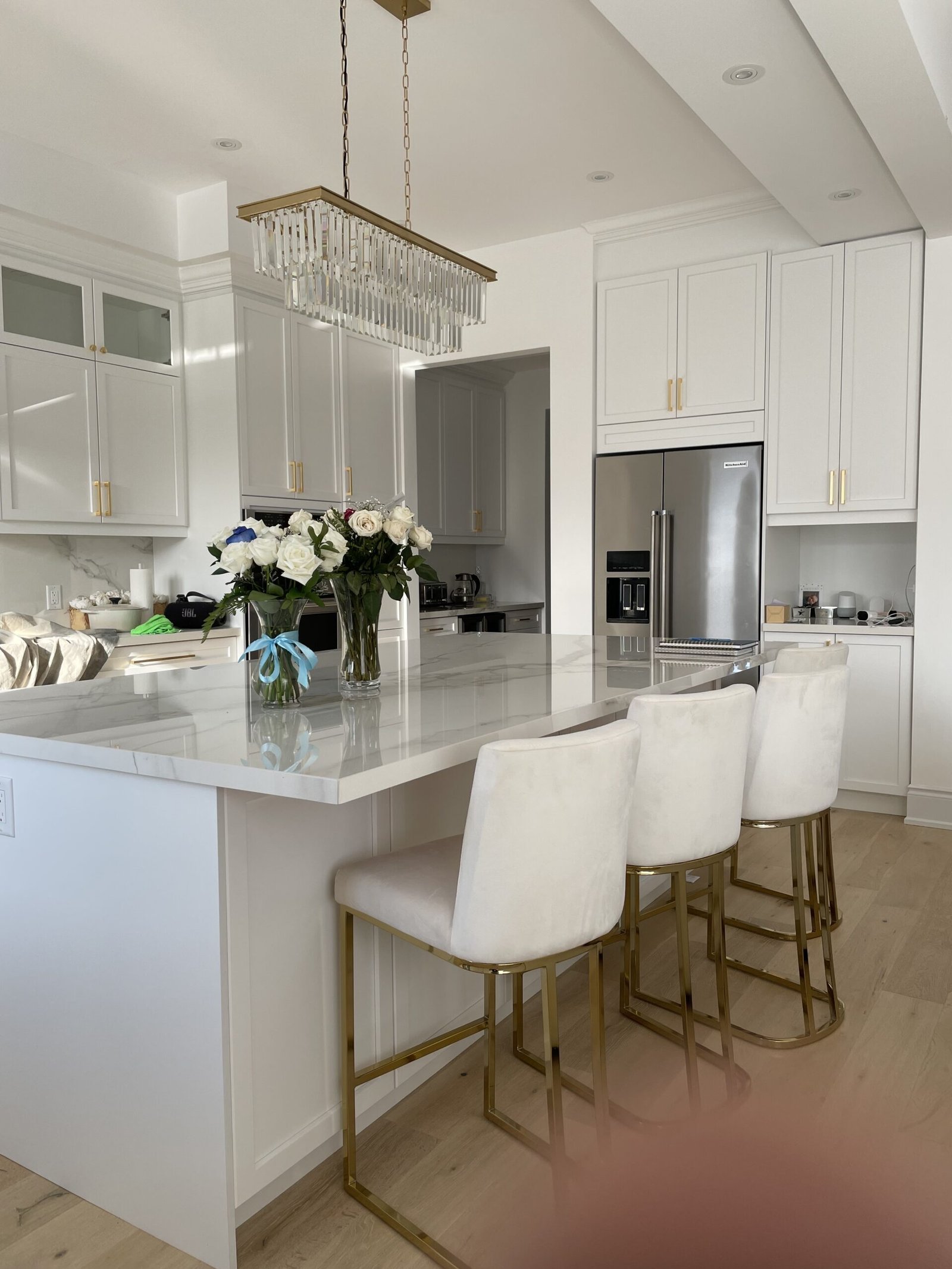 White kitchen with marble island, crystal chandelier, and gold-legged bar stools; bouquet of white roses on the counter nearby.