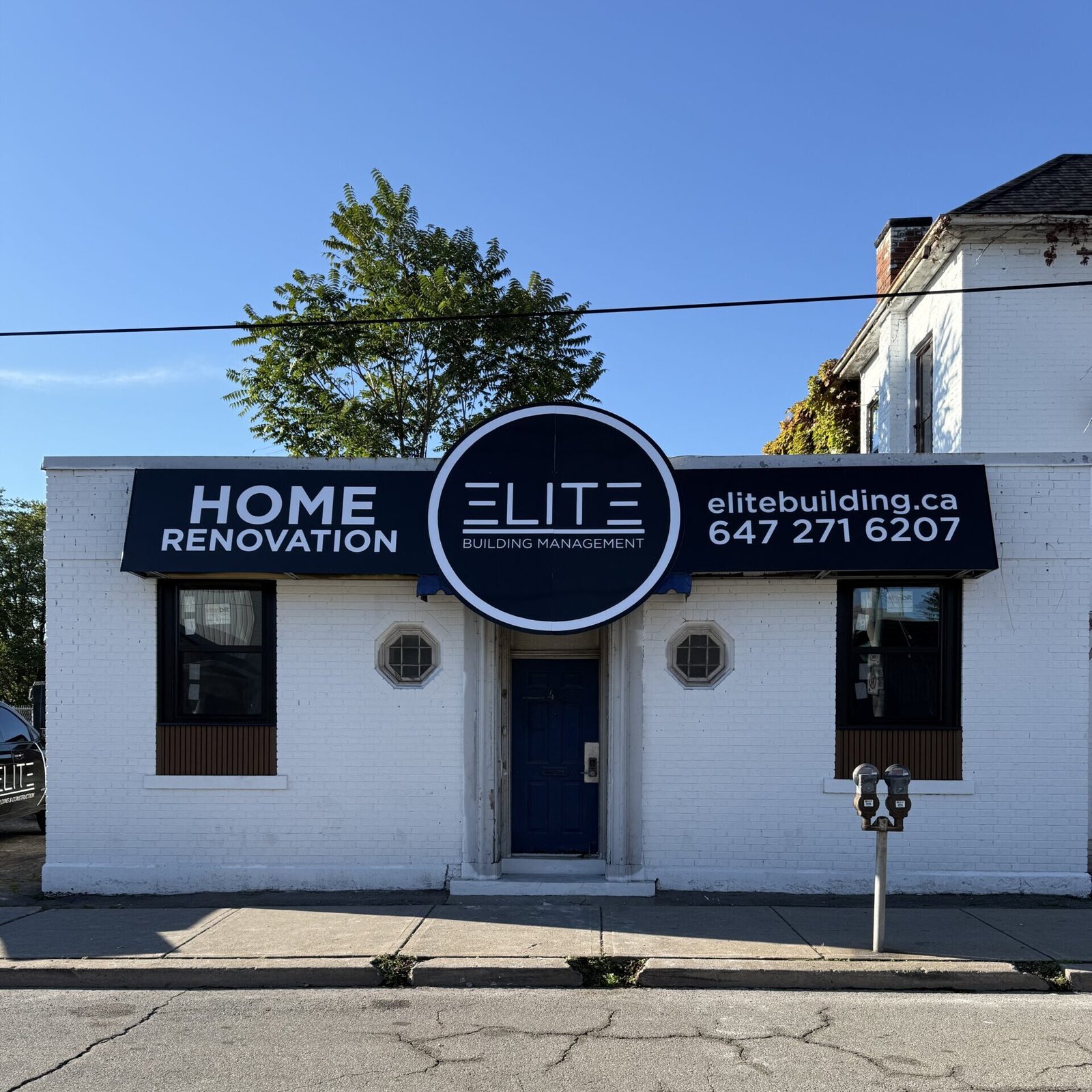White brick storefront with a large circular Elite logo sign and the words HOME RENOVATION on a dark blue panel, blue sky above, tree to the left.
