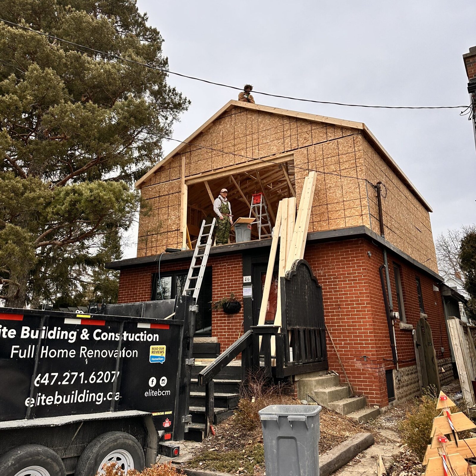Two construction workers stand on the roof of a partially framed brick building, with a construction trailer in the foreground.