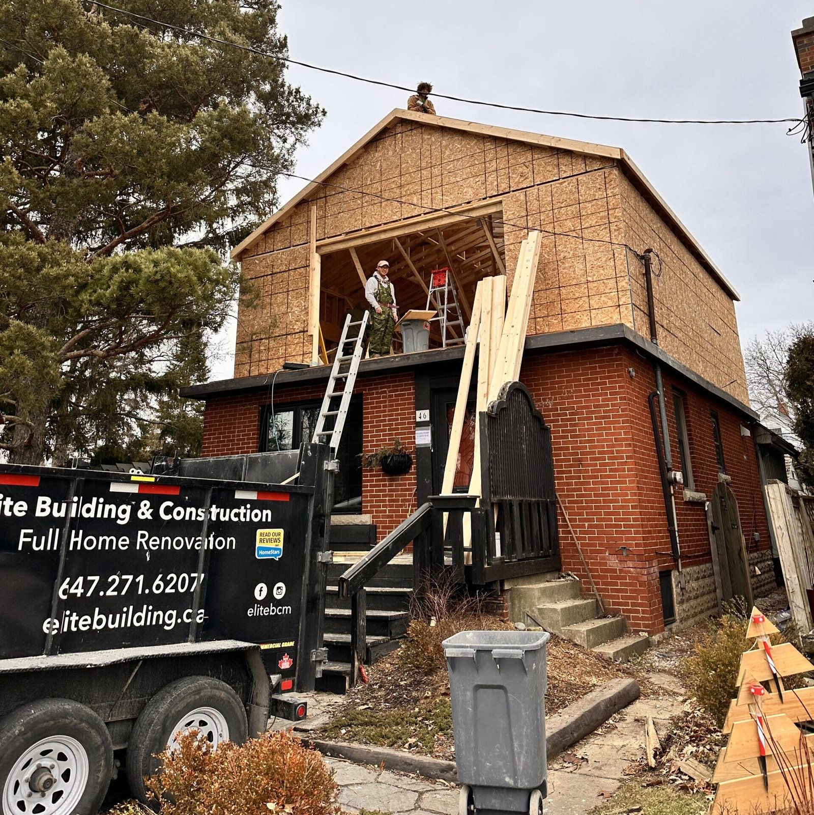 Two construction workers stand on the top edge of a brick building under renovation, with exposed plywood sheathing and lumber nearby.