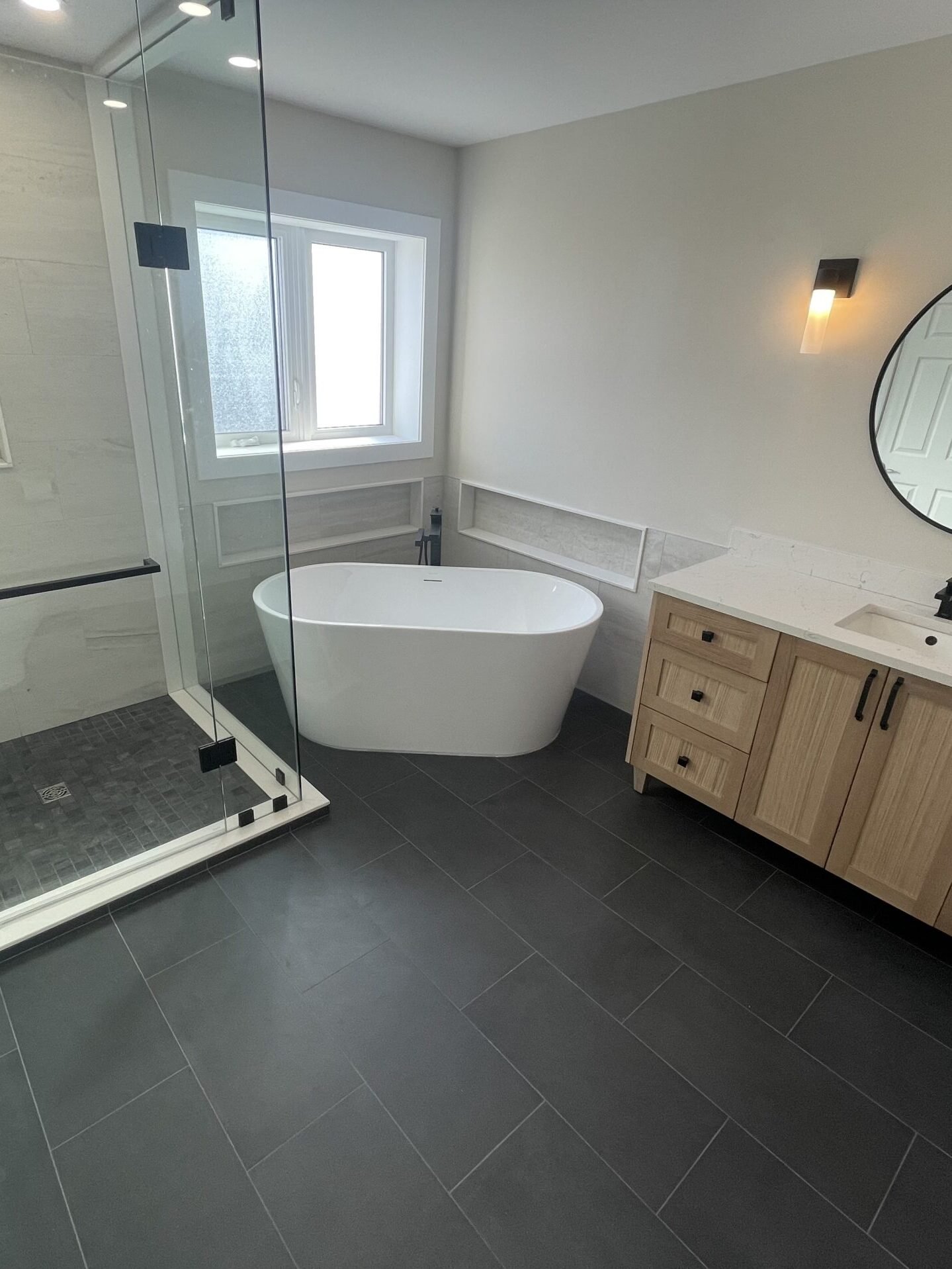 Modern bathroom with glass shower enclosure, freestanding white tub, and a wooden vanity under a round mirror on a neutral wall.