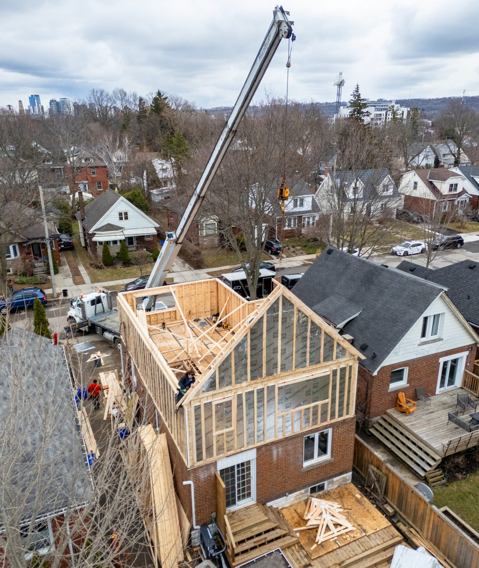 Crane lifts building materials over a partially framed two‑story house in a suburban neighborhood.