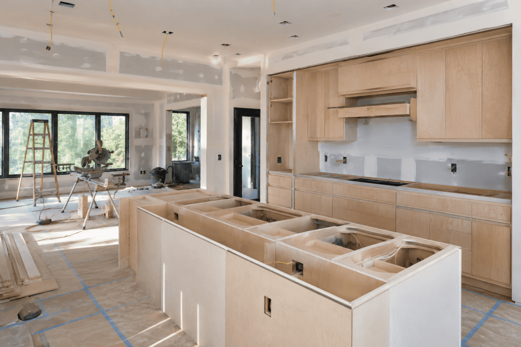 Kitchen renovation in progress: unfinished light wood cabinets, island frame, and exposed drywall with yellow wiring.